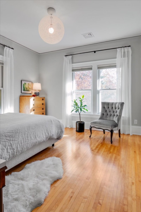 Bedroom with chair, bed, plant and dresser with small lamp and artwork