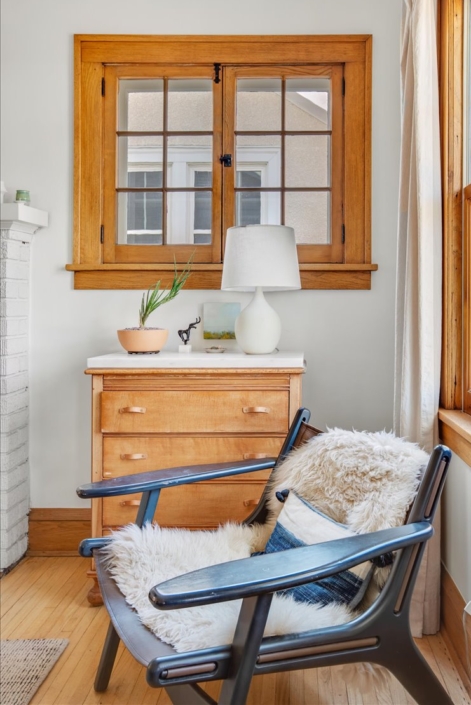 Cozy corner of family room with black chair covered by a sheepskin and a small refinished dresser with leather pulls, ceramic lamp and unique plant by small art